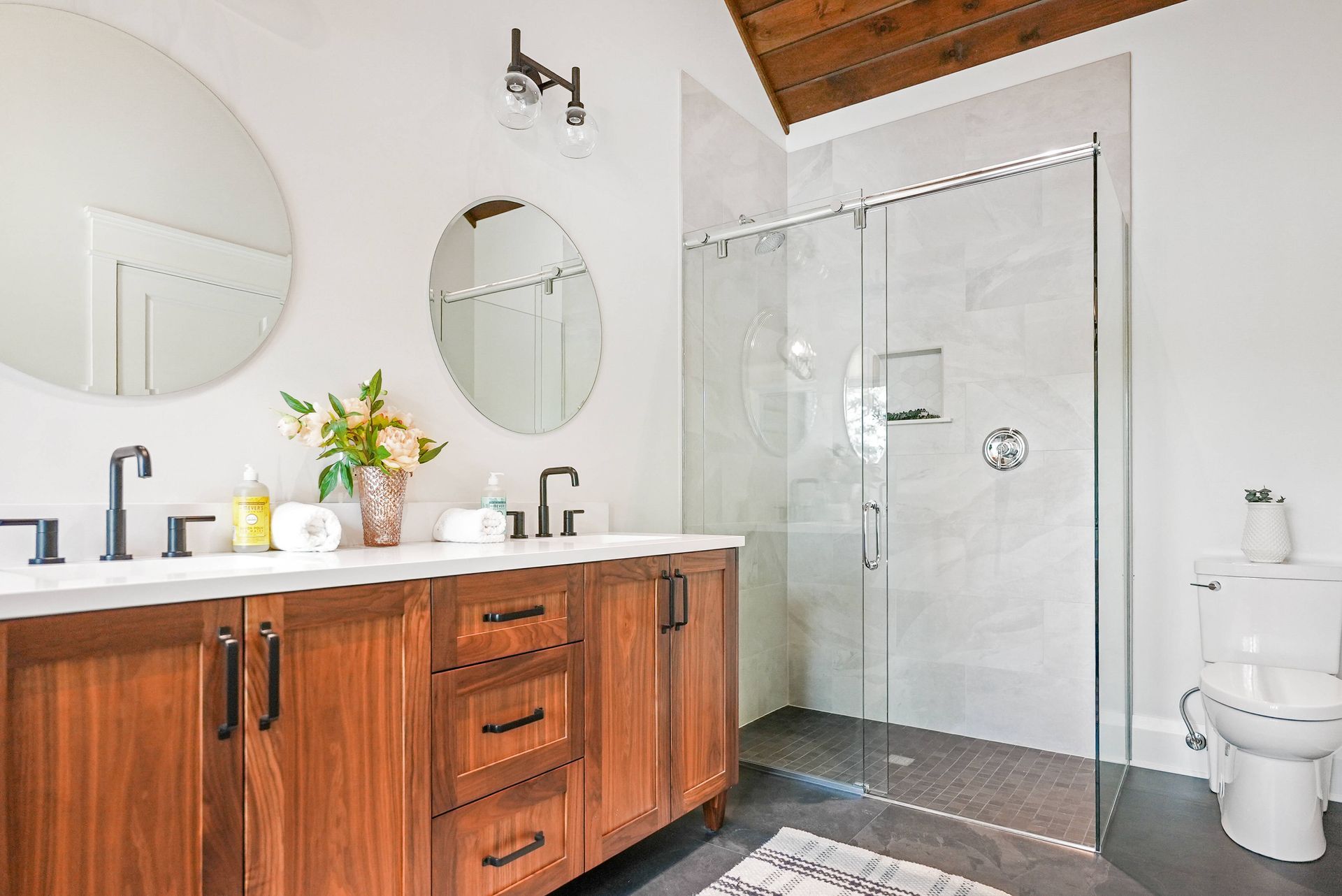 Modern bathroom with wood vanity, double sinks, round mirrors, glass shower, and a toilet