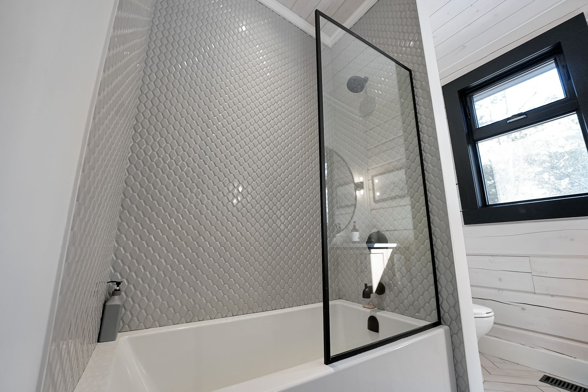 Modern bathroom with a glass shower door, gray mosaic tile, and a small window.