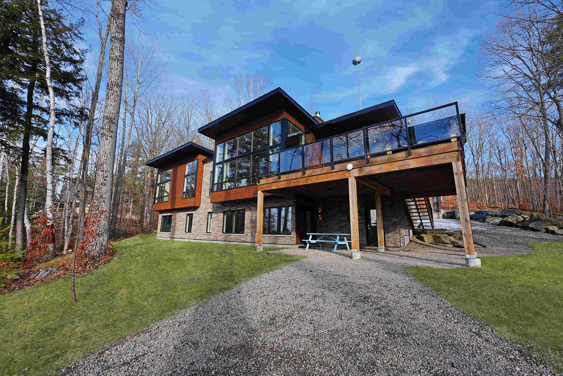 Modern two-story house with stone base, large windows, and a gravel driveway in a wooded area