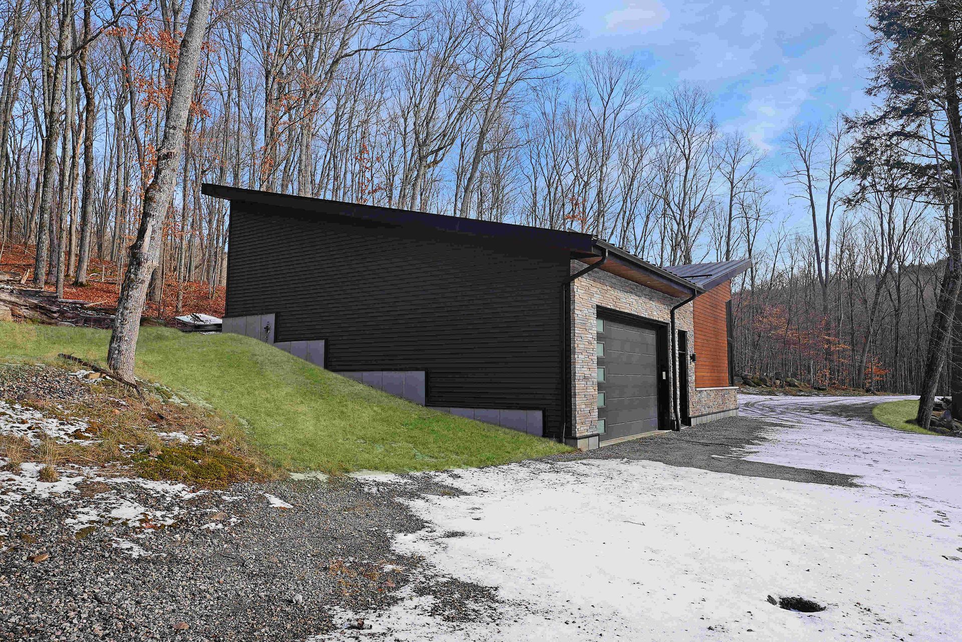 Modern black cabin in snowy woodland with leafless trees and a gravel driveway