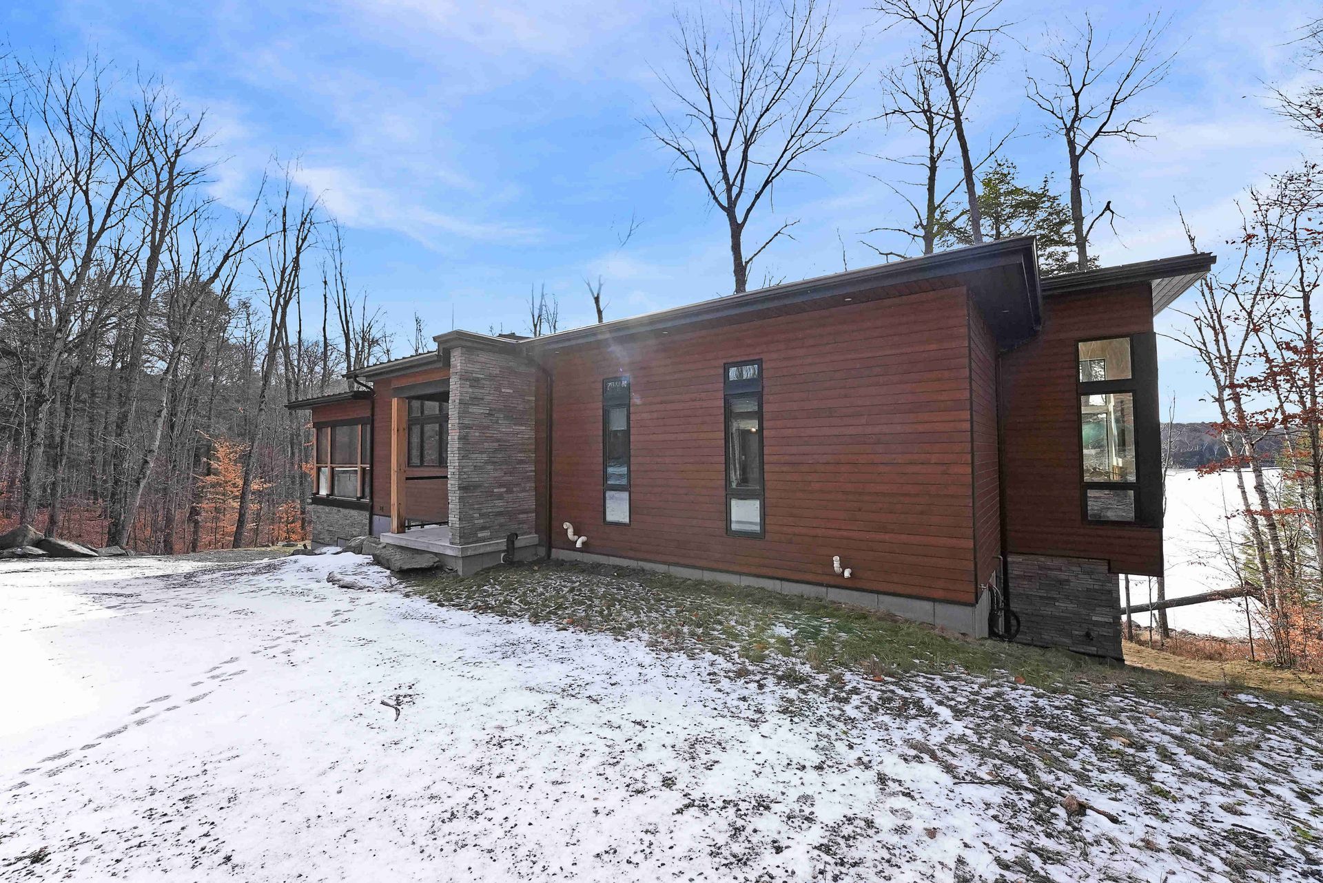 Snowy modern cabin with wood siding and large windows in a leafless wooded setting
