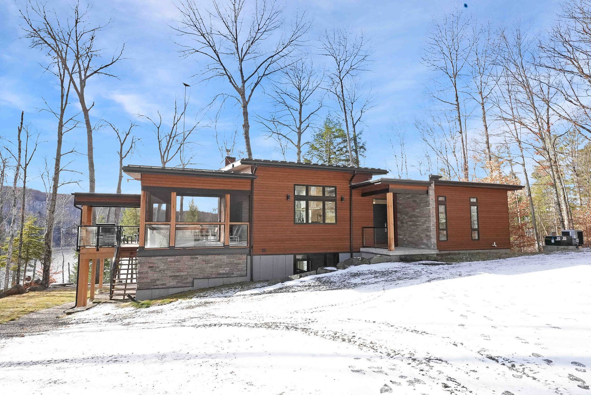 Modern wood cabin with large windows in a snowy, leafless forest under a clear blue sky