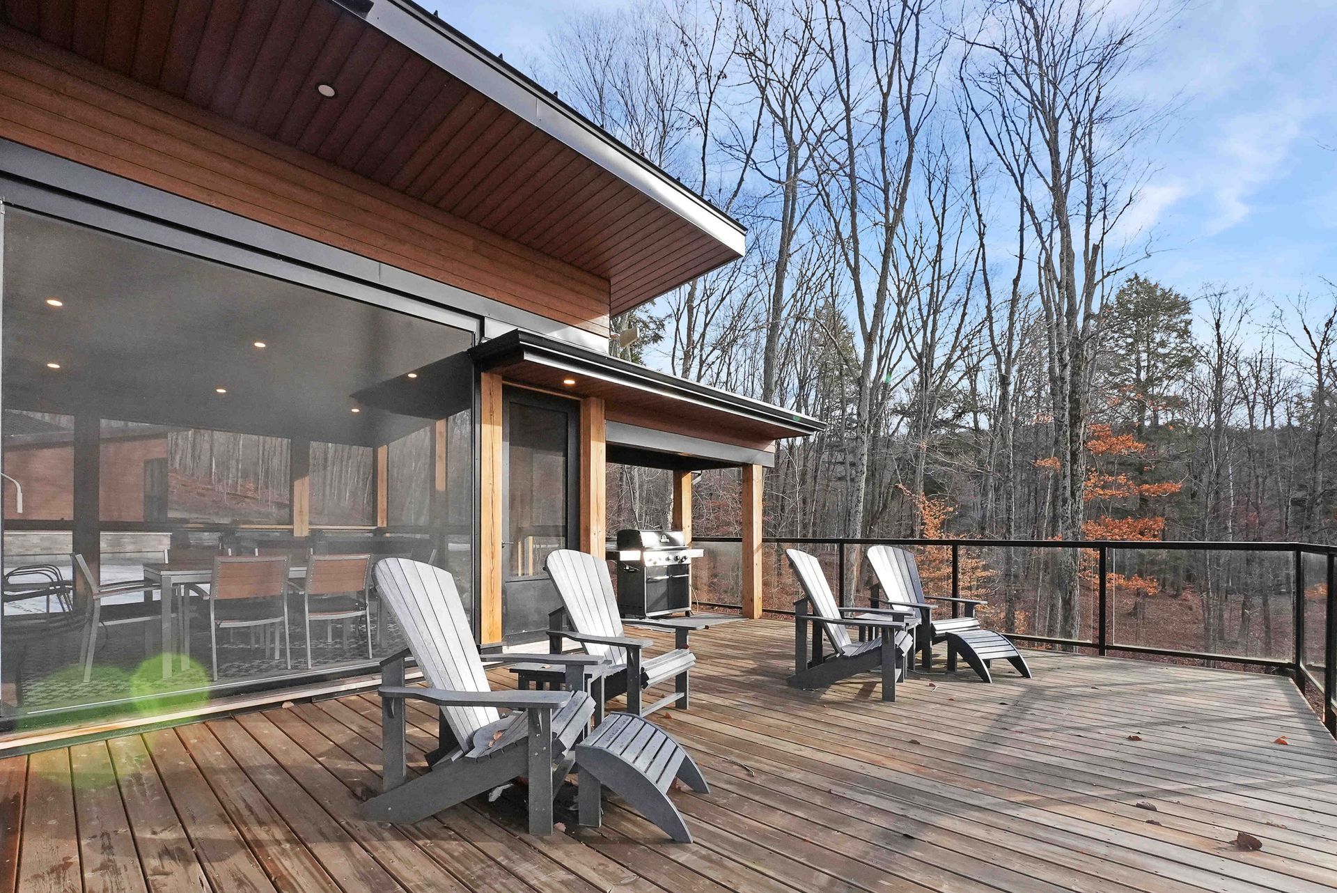 Wooden deck with lounge chairs beside a modern house, overlooking bare trees and a cloudy yard.