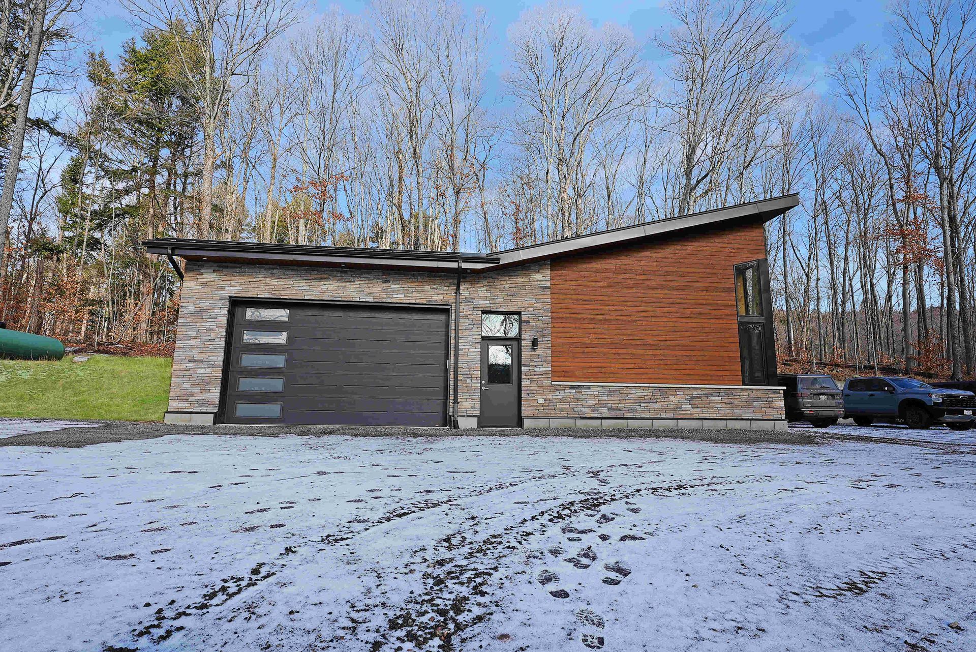 Modern house with garage, wood siding, and snow-covered ground in a wooded area