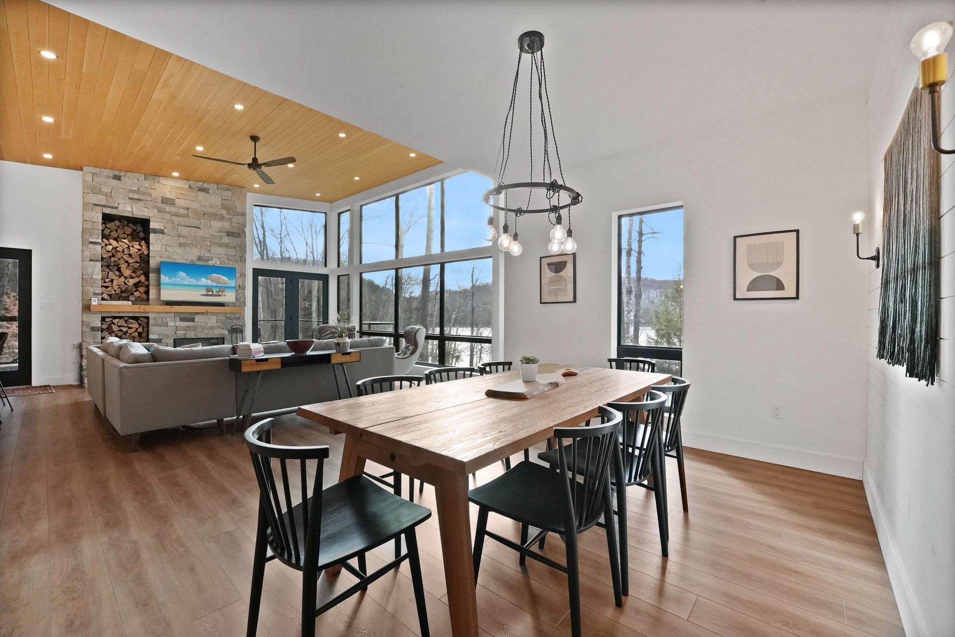 Bright dining room with long wooden table, black chairs, high ceiling, and large windows overlooking a lake.