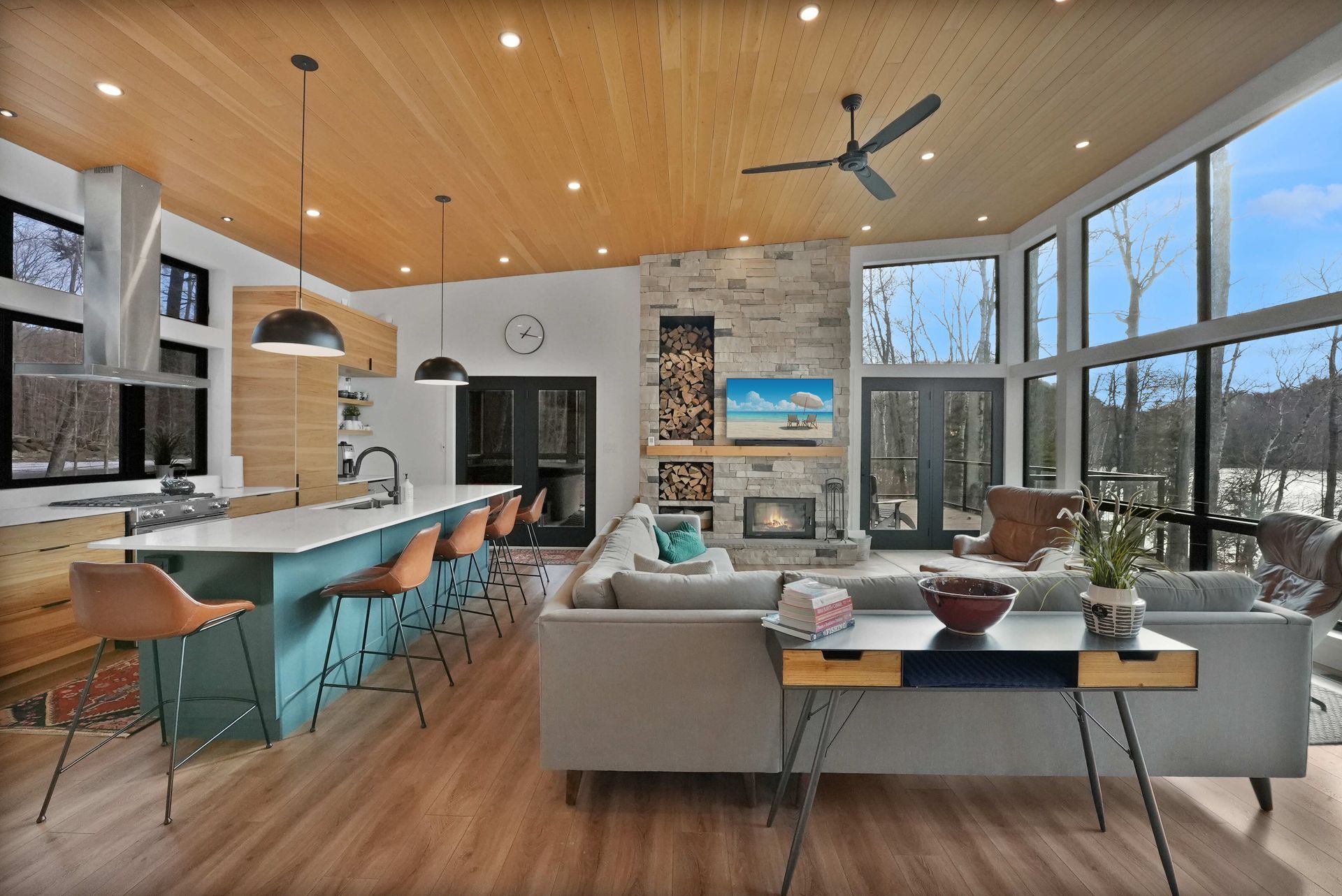 Modern open-plan living room with kitchen island, gray sofa, wood ceiling, and large windows overlooking a lake.