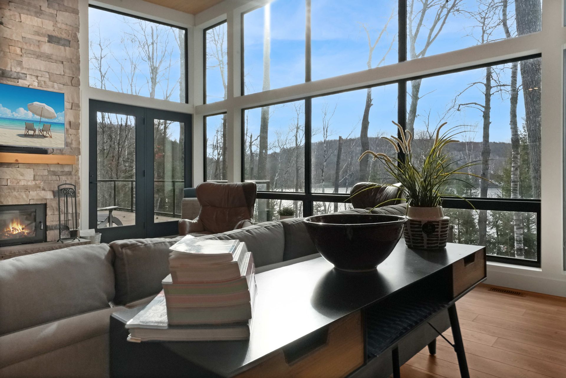 Sunlit modern living room with a gray sofa, black table, fireplace, and tall windows overlooking trees