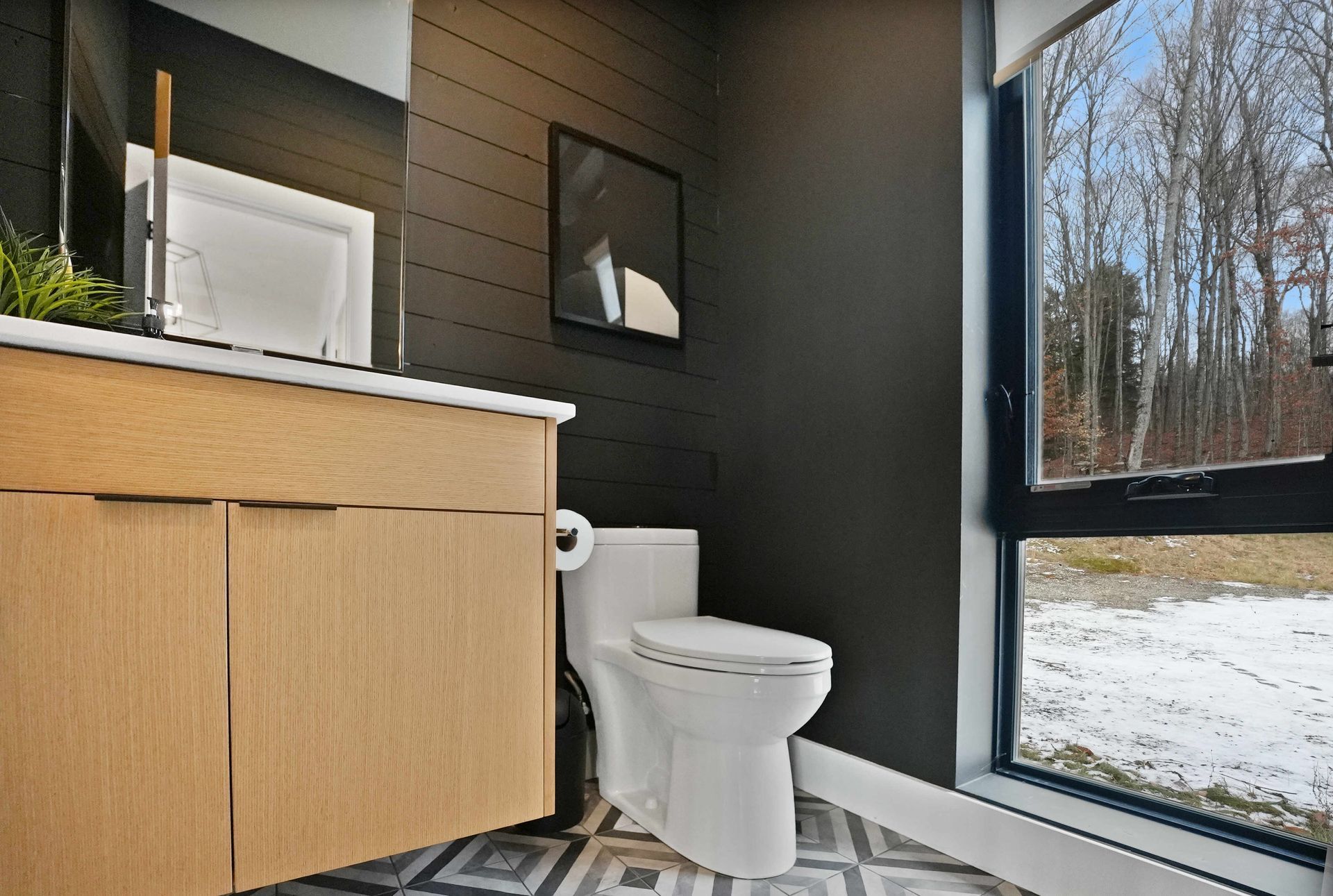 Modern bathroom with wood vanity, toilet, dark walls, and a large window showing snowy trees.