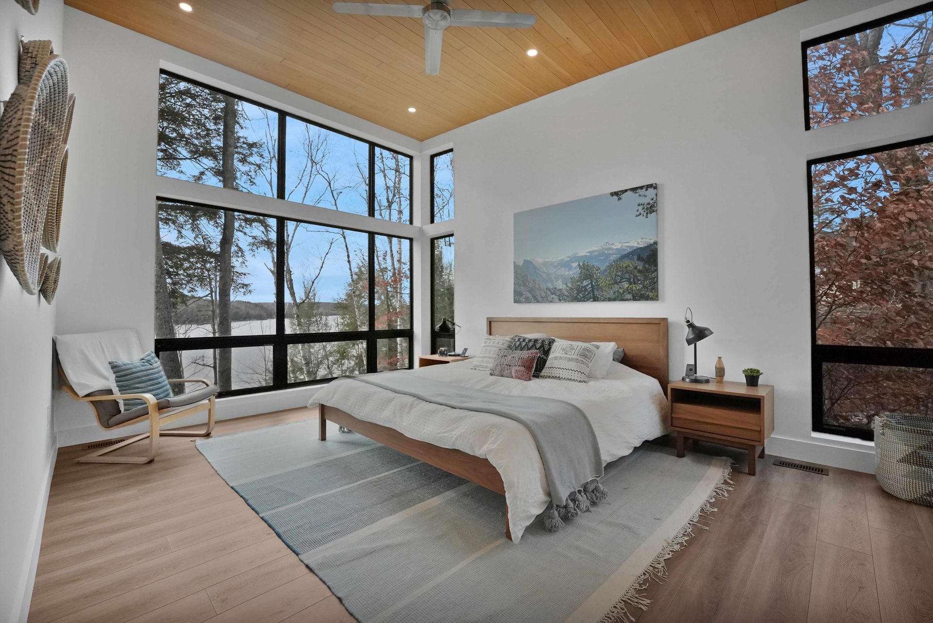 Modern bedroom with floor-to-ceiling windows, wood ceiling, and a bed with gray bedding and a landscape print above it