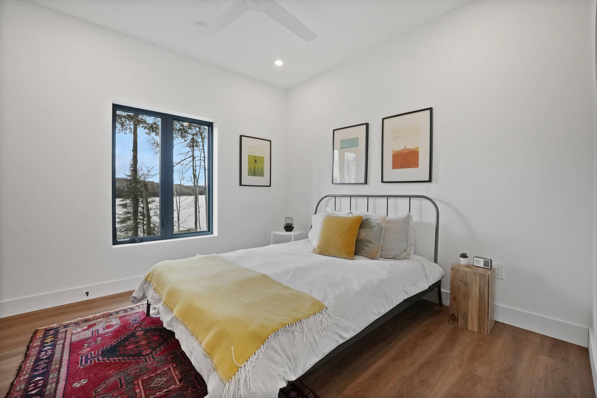 Bright bedroom with white walls, metal bed, yellow bedding, framed art, and a window with trees outside