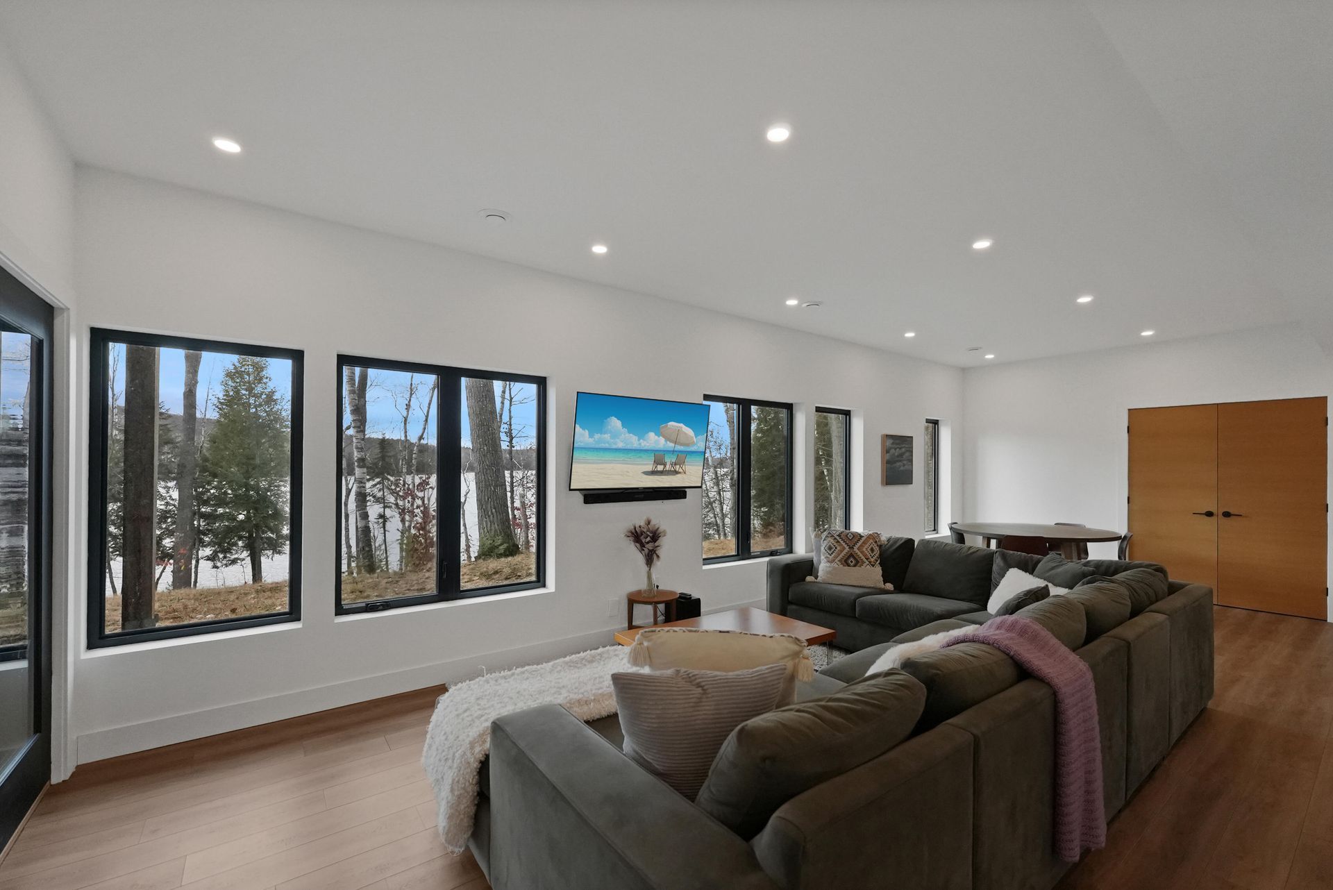 Bright modern living room with large windows, gray sectional sofa, wall-mounted TV, and hardwood floors
