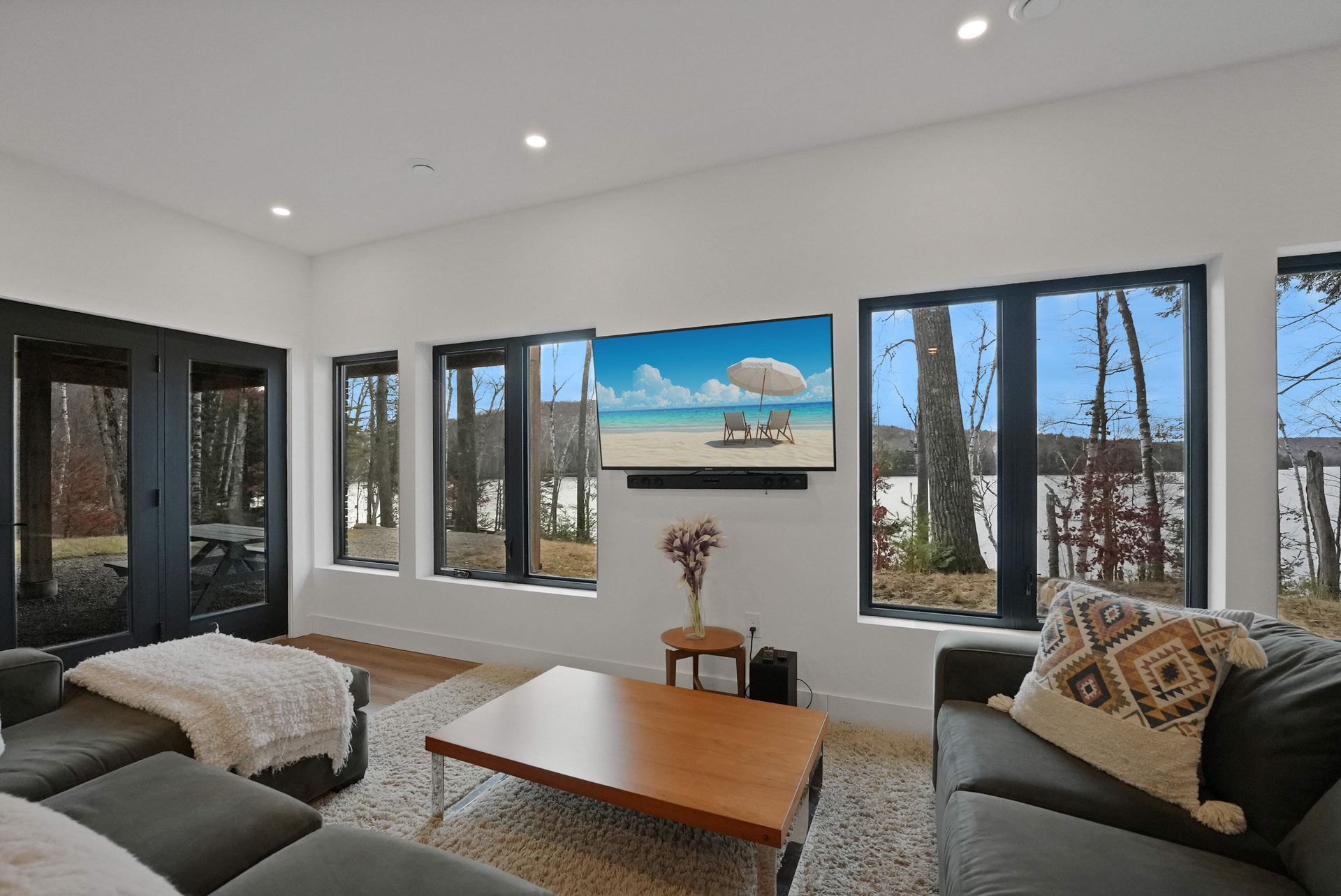 Bright living room with large windows, gray sofas, and a wall-mounted TV above a wooden table