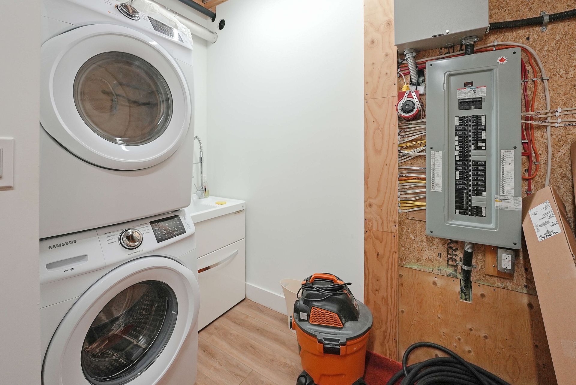Laundry room with stacked washer and dryer beside an open electrical panel on unfinished wall