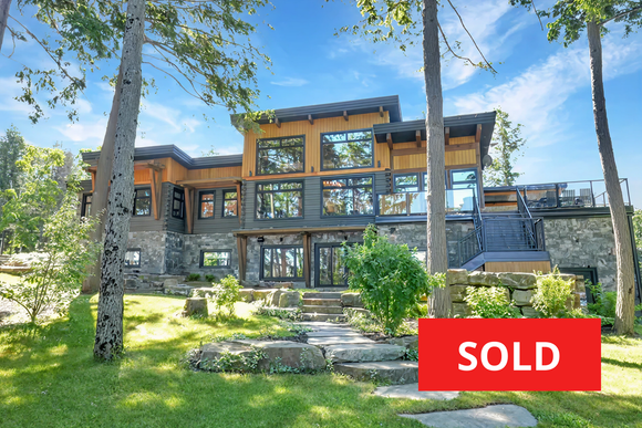 Modern stone-and-wood lake house with large windows, surrounded by trees; red SOLD sign in front.