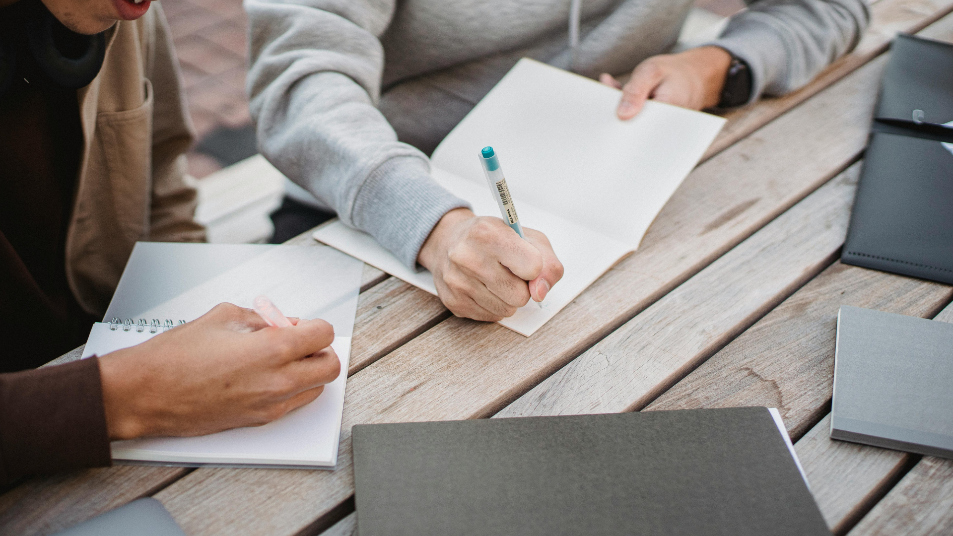 Hands writing on paper at a wooden table with notebooks and a laptop nearby