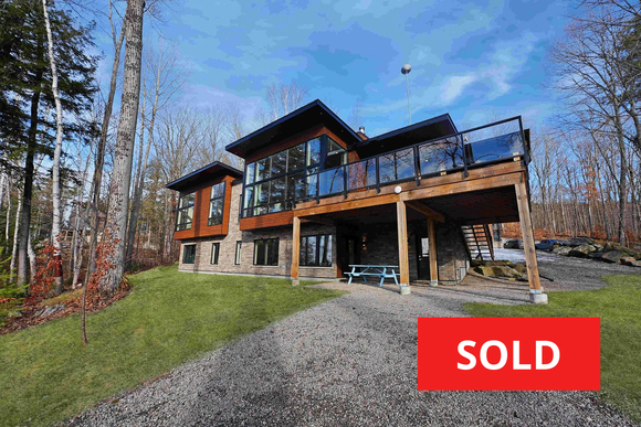 Modern two-story house with deck and gravel driveway, marked with a red “SOLD” sign.