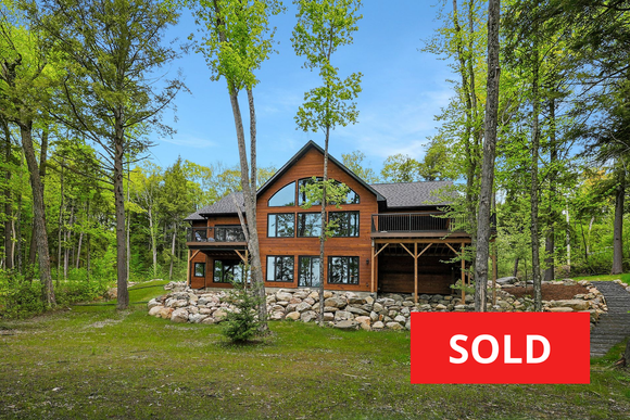 Wooden cabin with stone foundation in a sunny forest clearing, surrounded by tall green trees.