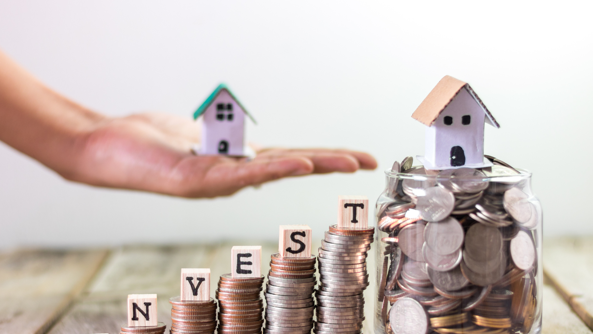 A hand holds a small model house above coin stacks spelling