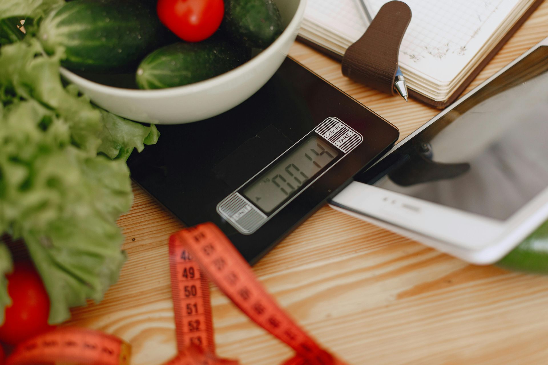 Kitchen scale with fresh vegetables, measuring tape, and a notebook on a wooden table