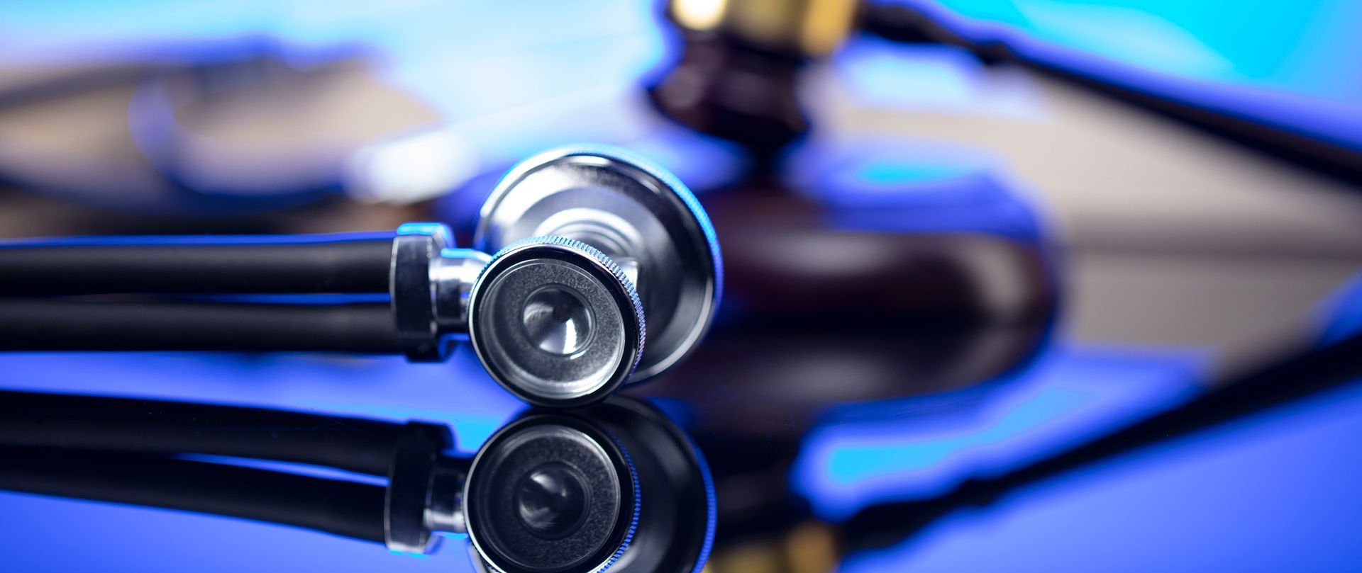 Stethoscope resting on a reflective surface with a blurred gavel in the background. Blue lighting.