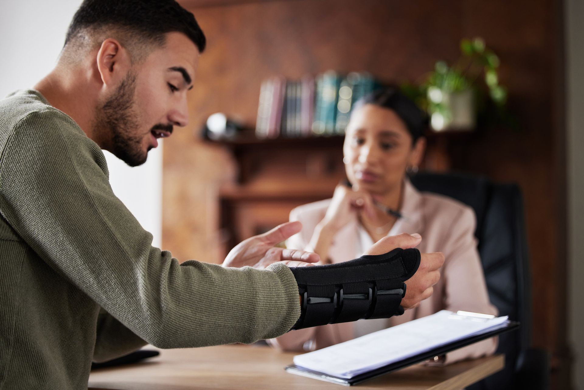 Man in wrist brace gestures, speaking to a person at a desk holding a notepad. Interior setting.