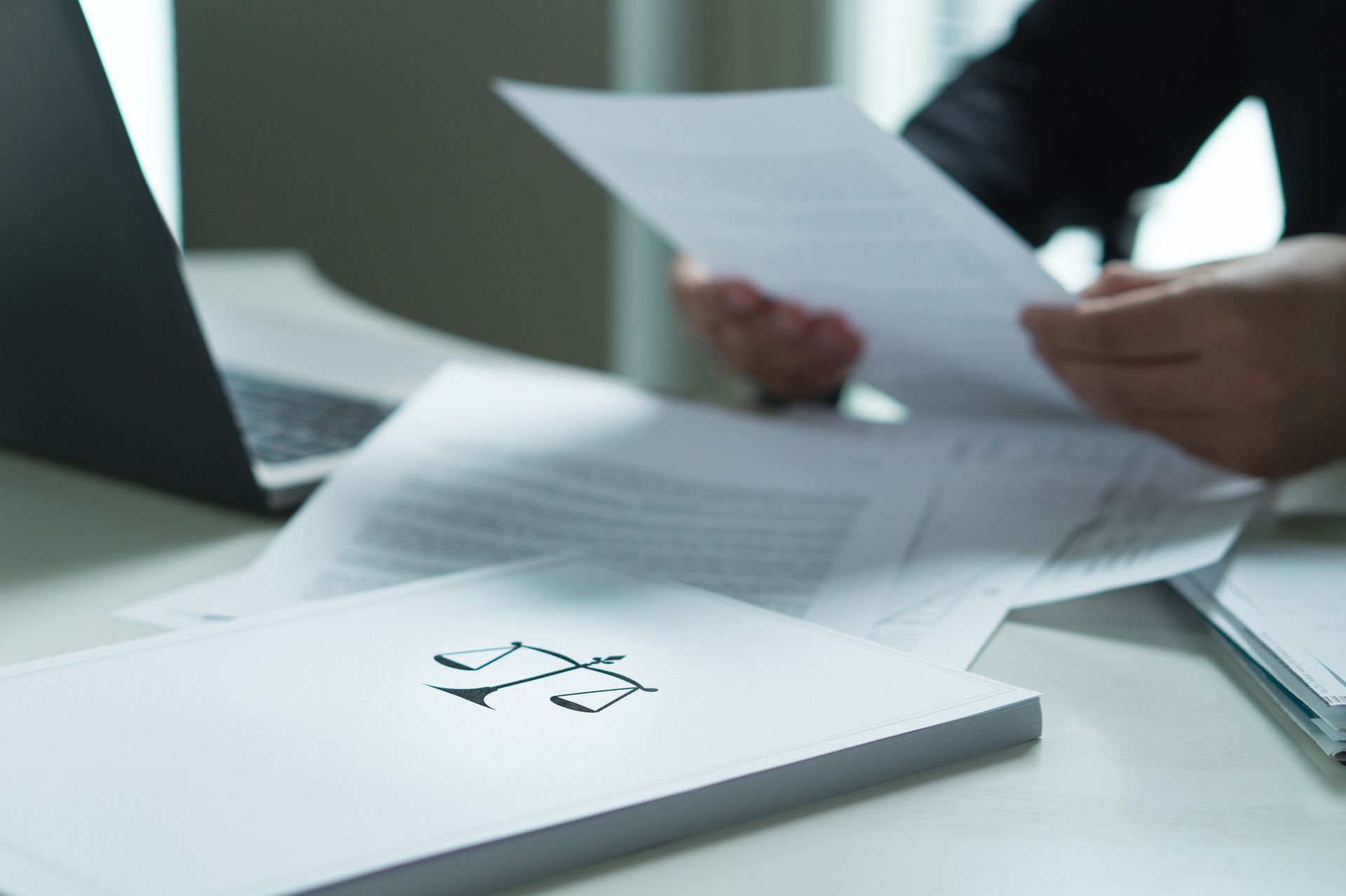 Professional reviewing legal documents at desk with laptop and justice scales folder visible.