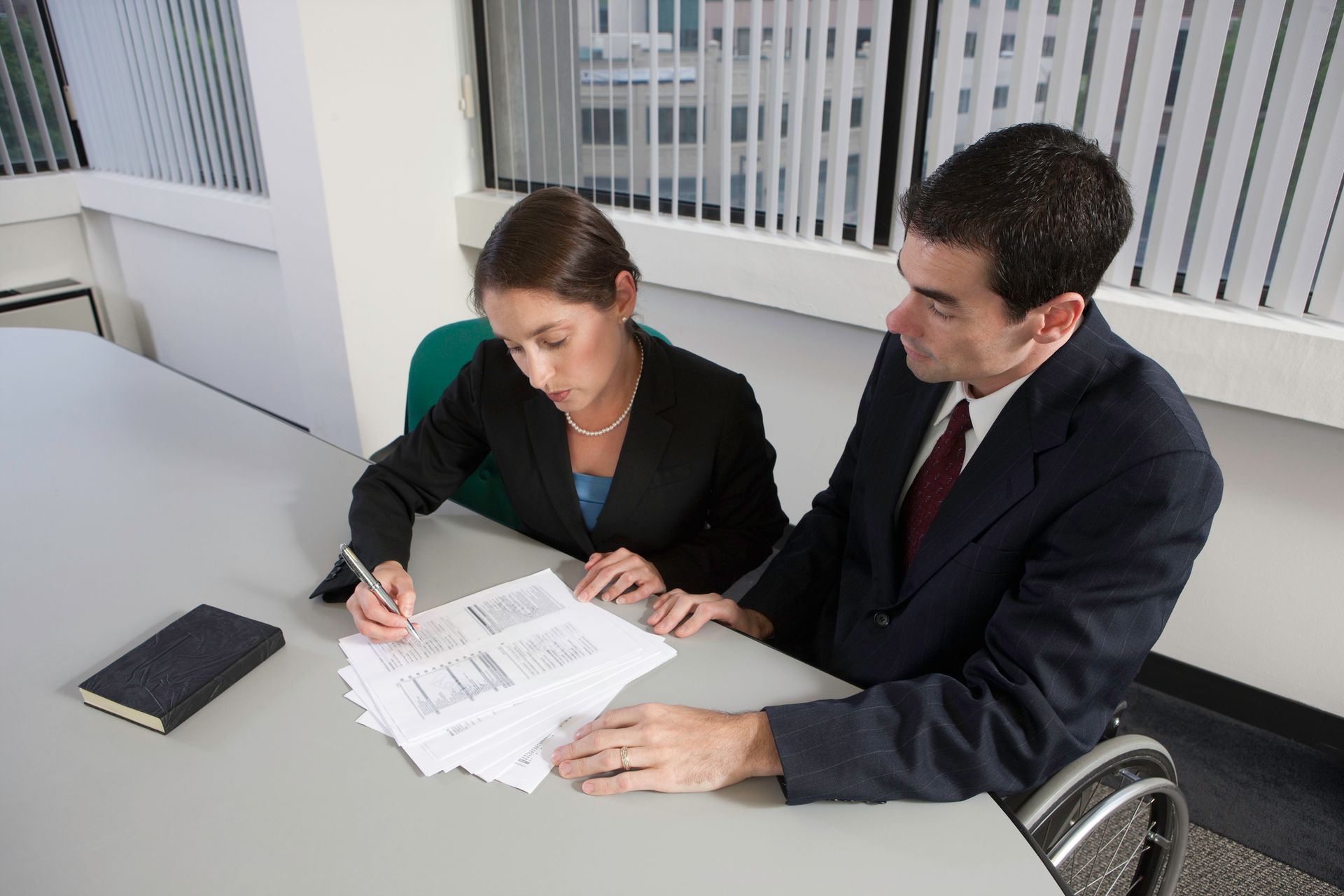 Woman and man reviewing documents at a table. Man in suit points, woman in suit writes. Inside office setting.