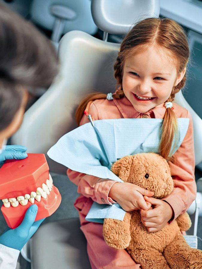 A little girl is sitting in a dental chair holding a teddy bear and a model of teeth.