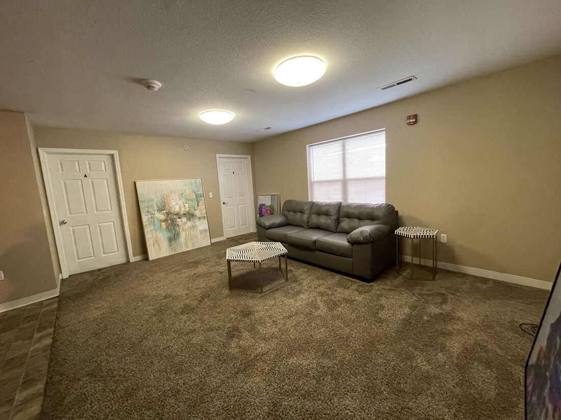 Living room with sofa, coffee table, and large artwork leaning against a door on a carpeted floor.