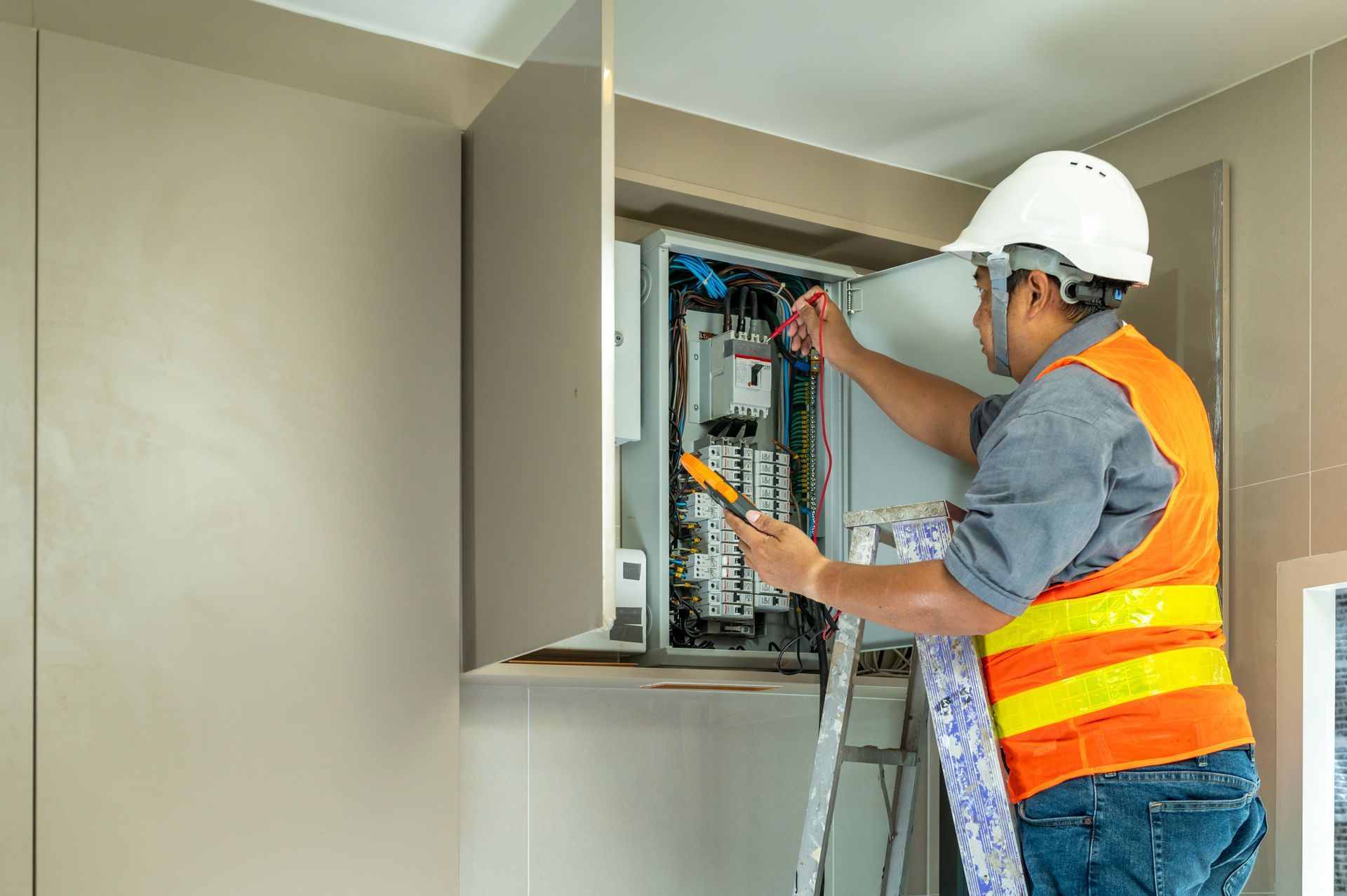 Electrician working on electrical panel, wearing a safety vest and helmet, standing on a ladder. Electrician working on electrical panel, wearing a safety vest and helmet, standing on a ladder.