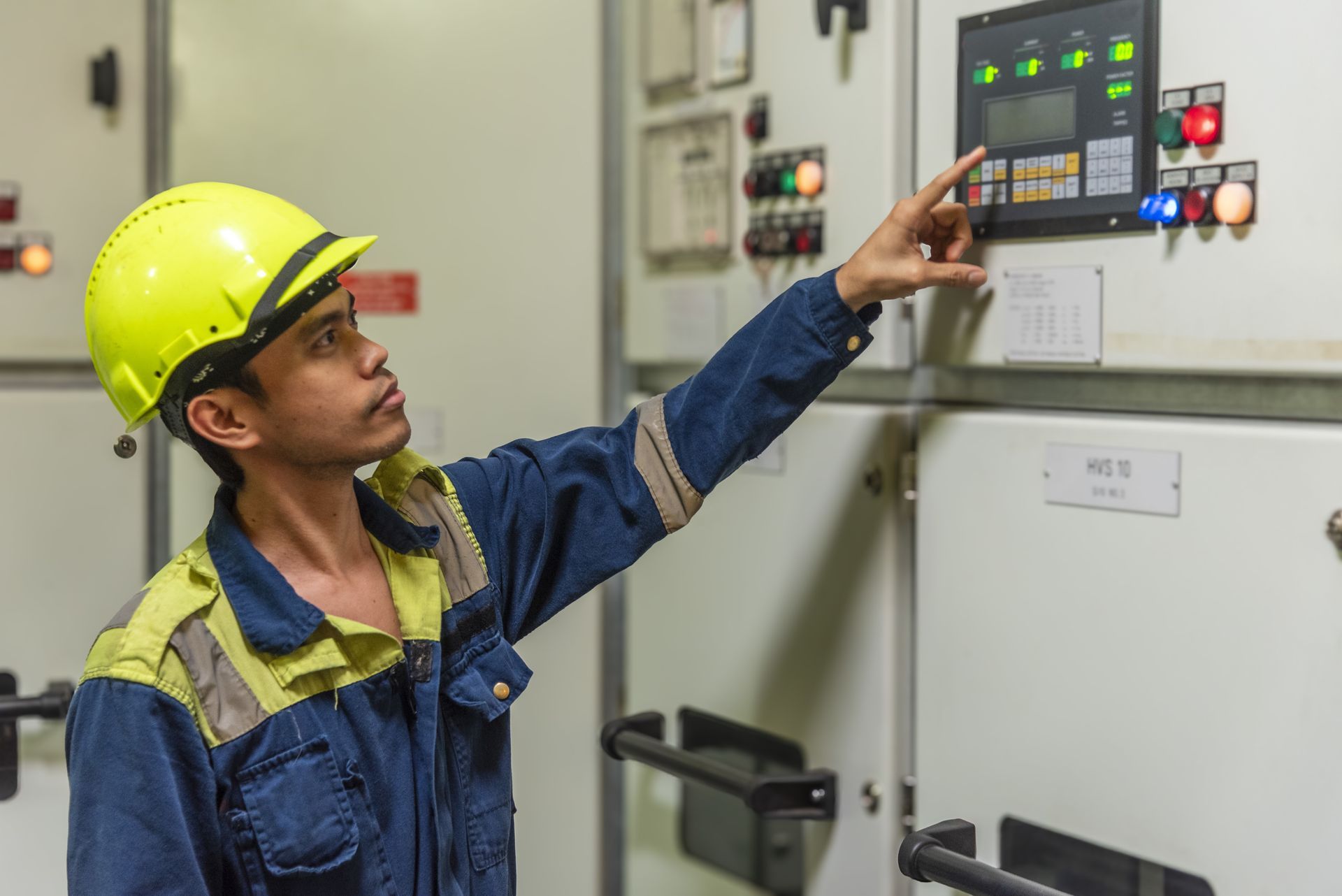 Technician in yellow hard hat and blue work suit inspecting electrical panel.