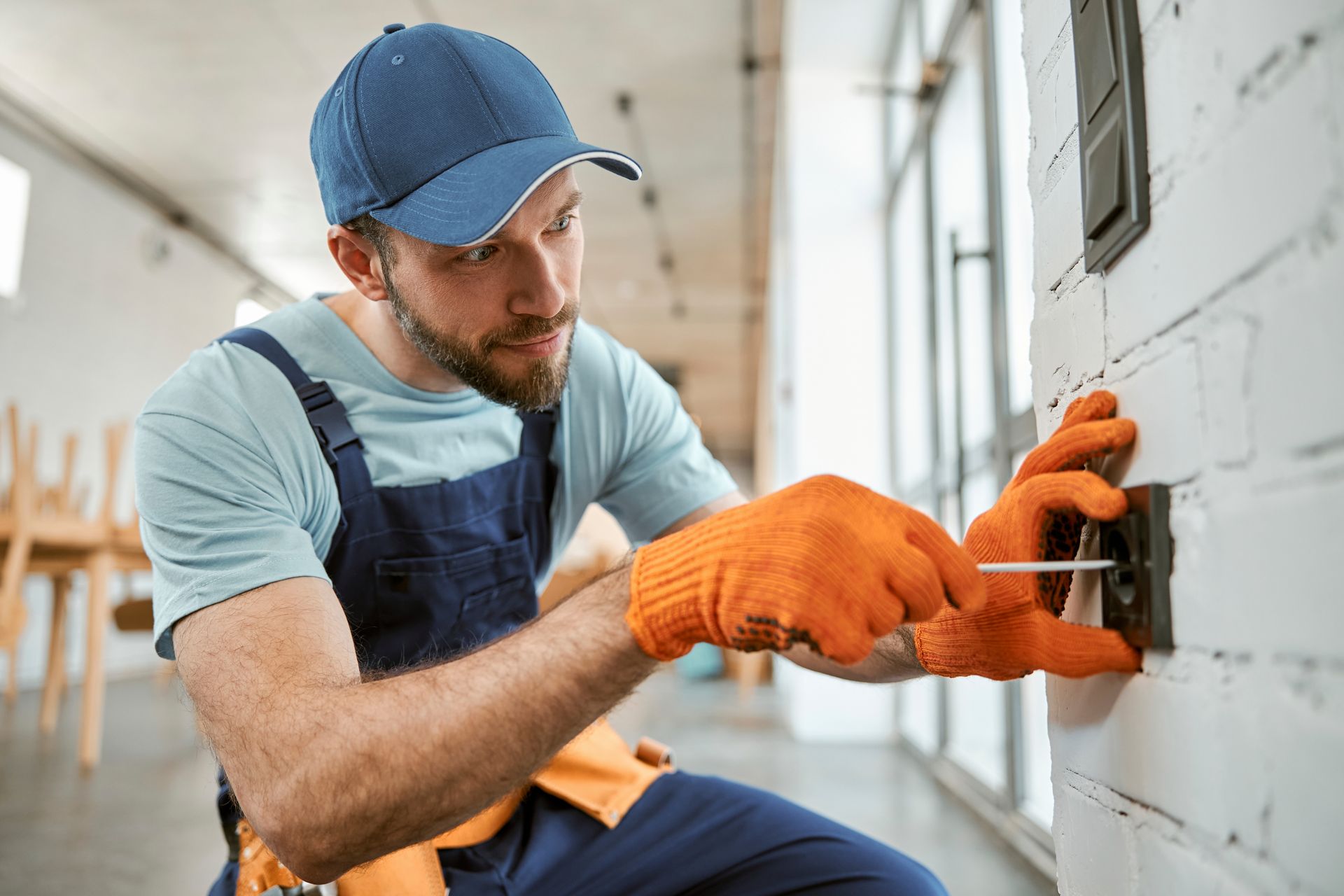 Electrician in orange gloves using a screwdriver on a wall switch. Brick wall, indoors. Electrician in orange gloves using a screwdriver on a wall switch. Brick wall, indoors.