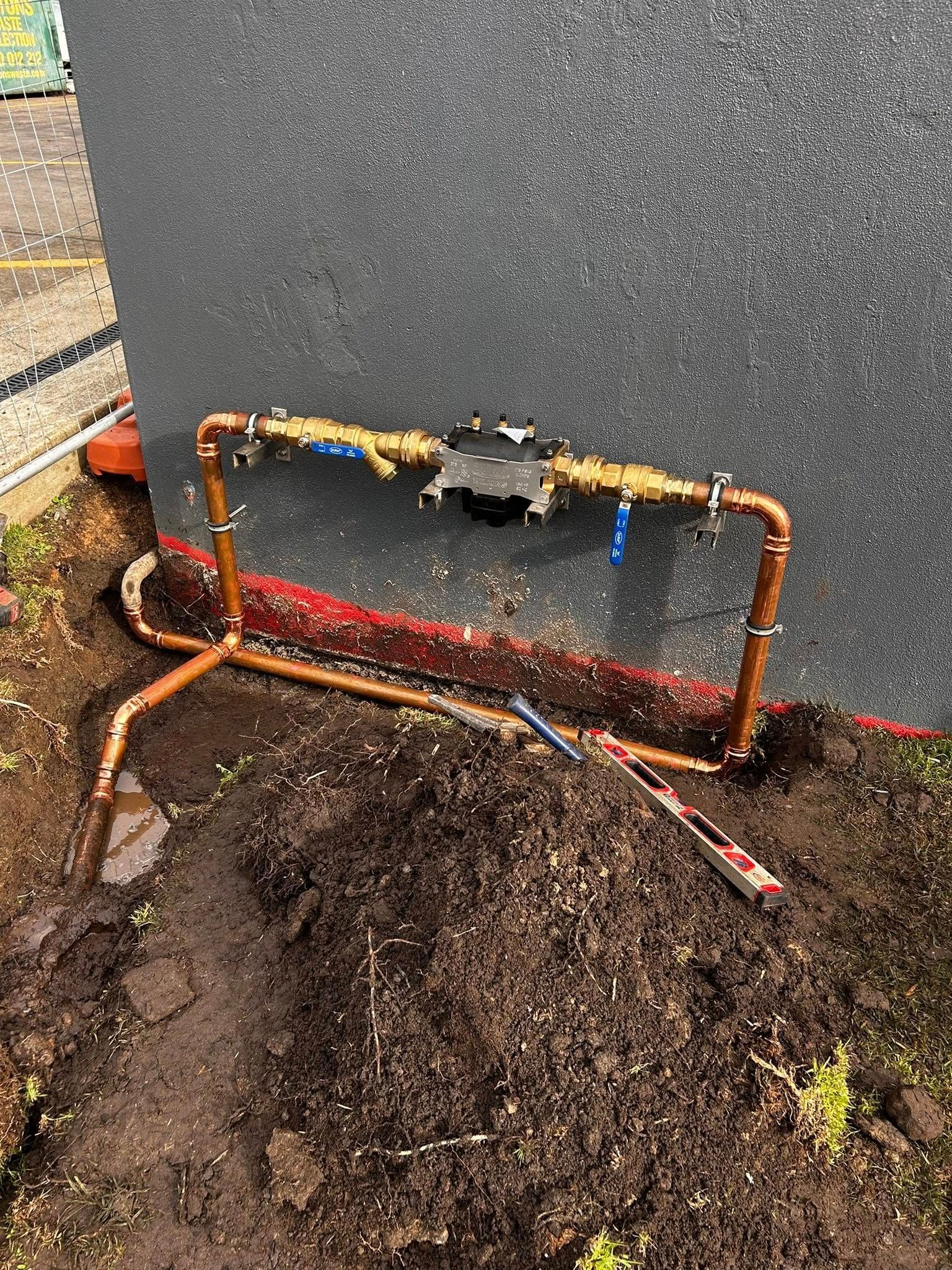 Copper pipes installed near a wall, in dirt.  A black device is in the pipes. Blue and yellow valves are attached.