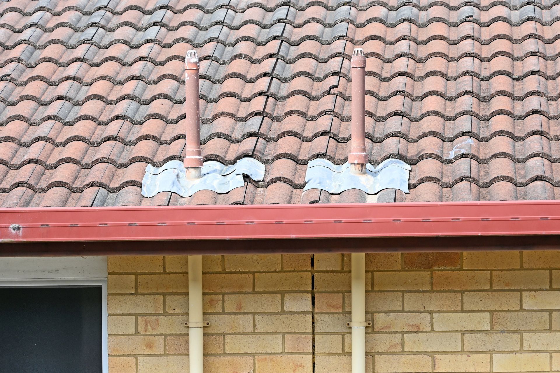 Brown tiled roof with two brown vent pipes, sealed with silver flashing, above a brick wall.