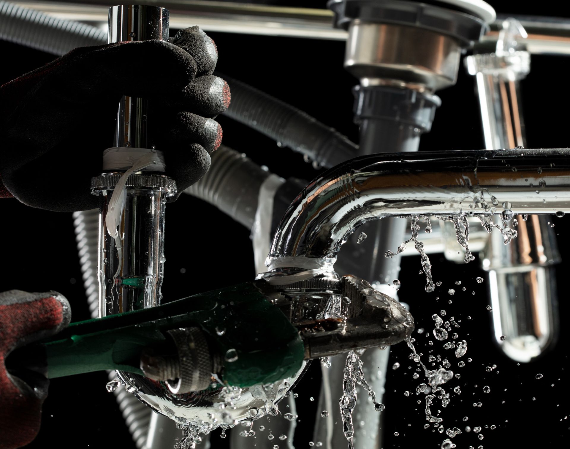 Plumber working on pipes under a sink; water spraying, wearing black glove and holding a wrench.