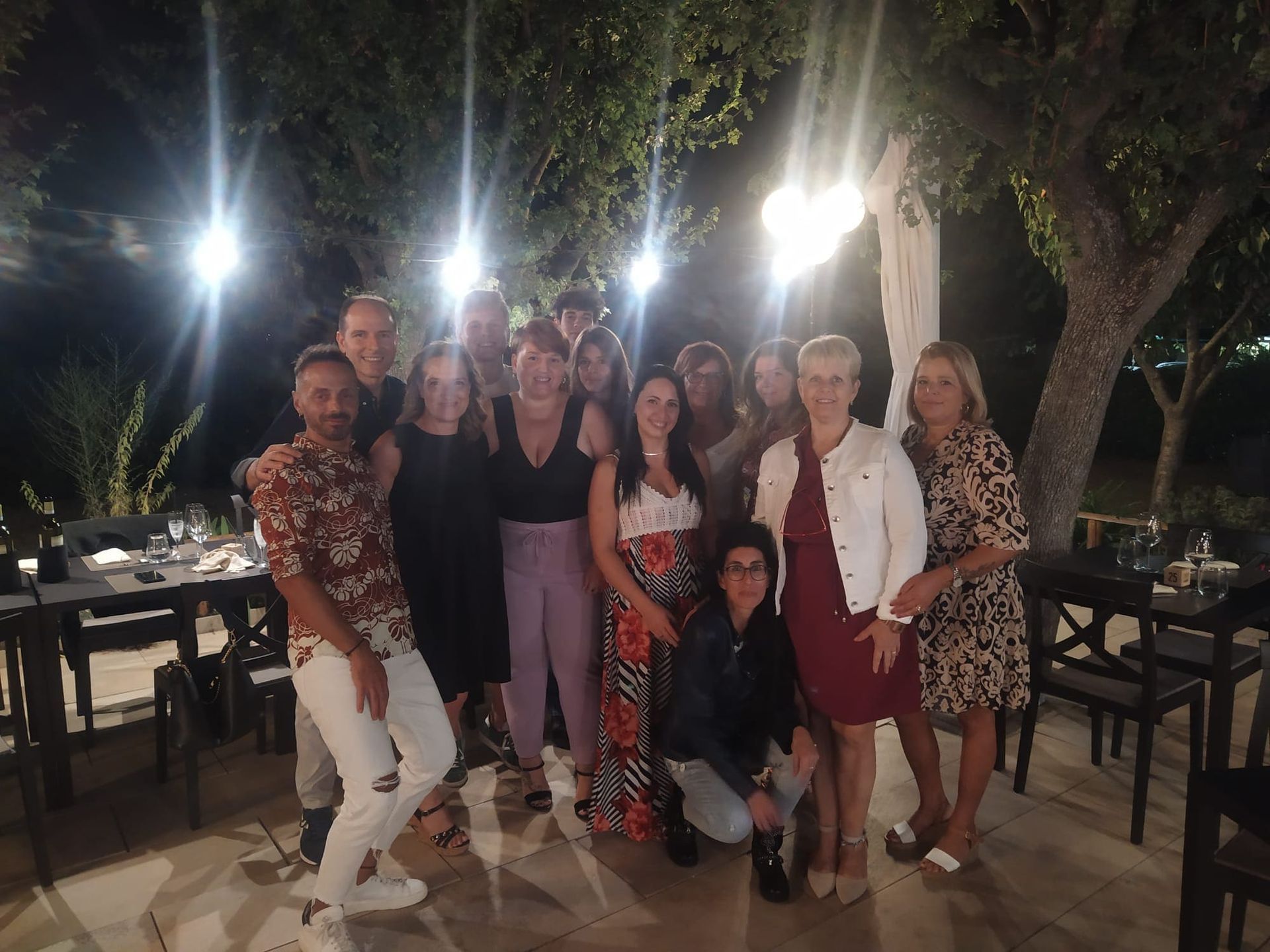 Gruppo di persone in posa all'aperto di notte su un patio illuminato con luci decorative e tavoli.