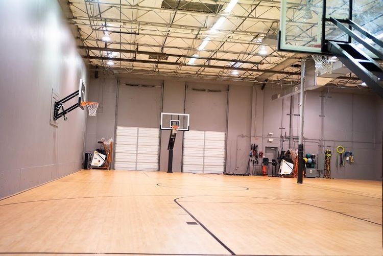 Empty indoor basketball court, hardwood floor, three hoops, gray walls, and a high ceiling.