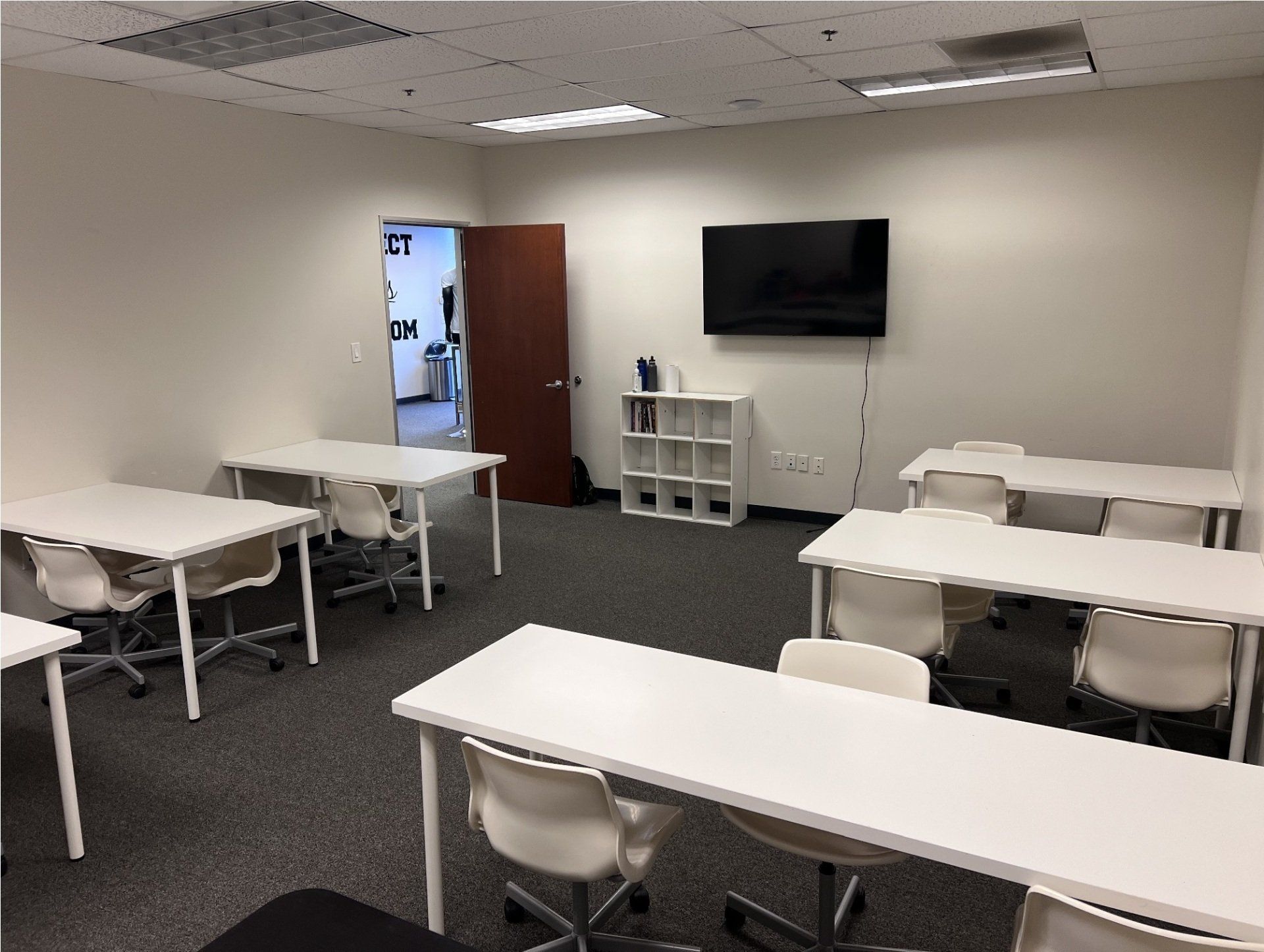 Empty classroom with white desks, chairs, and a mounted TV. Door open to a hallway.