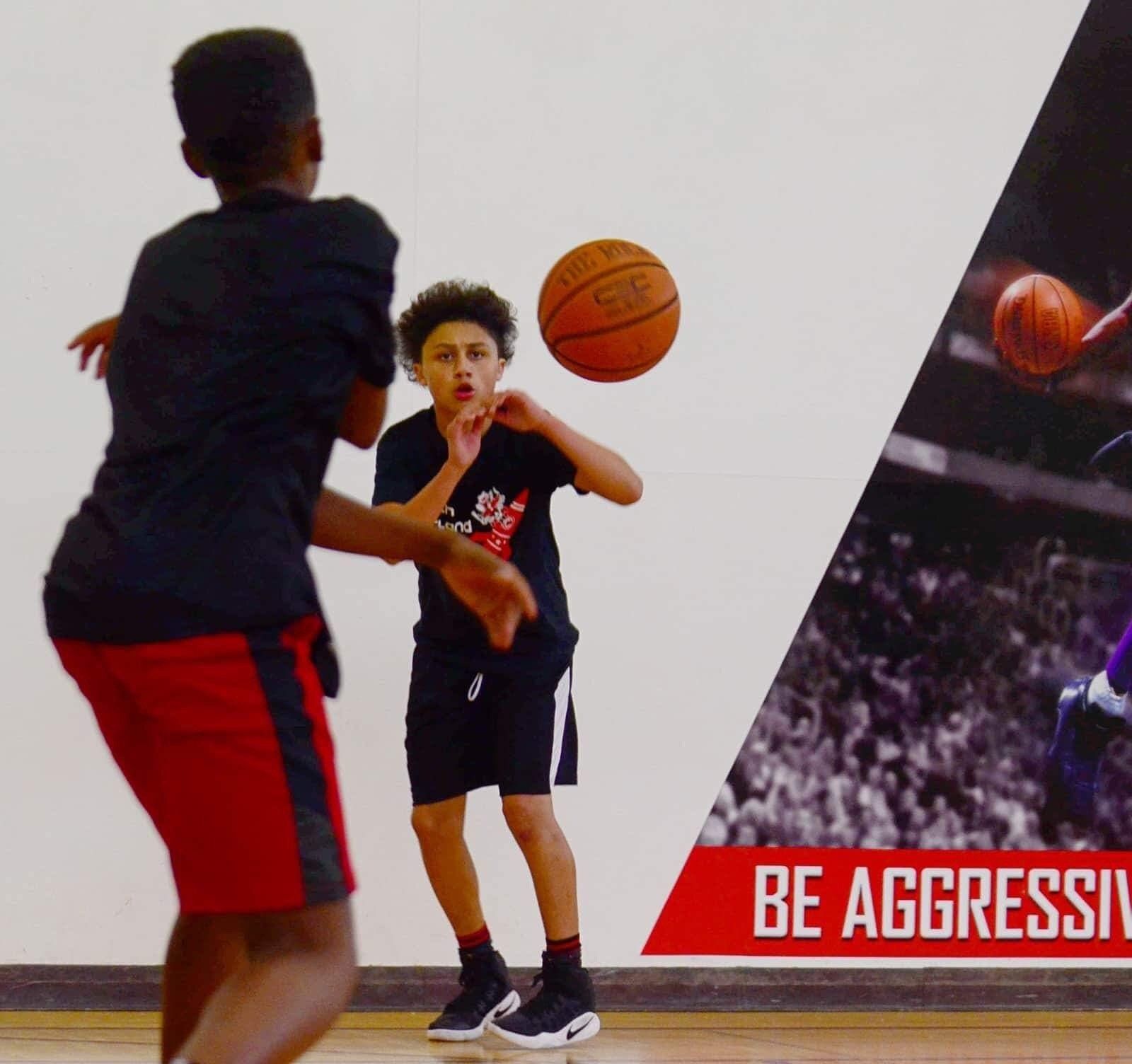 Two young boys playing basketball indoors; one about to pass the ball.