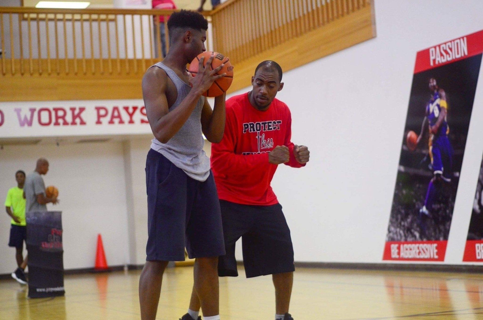 Basketball coach instructs a young player, in a gym. Red shirt, dark shorts, holding the ball.