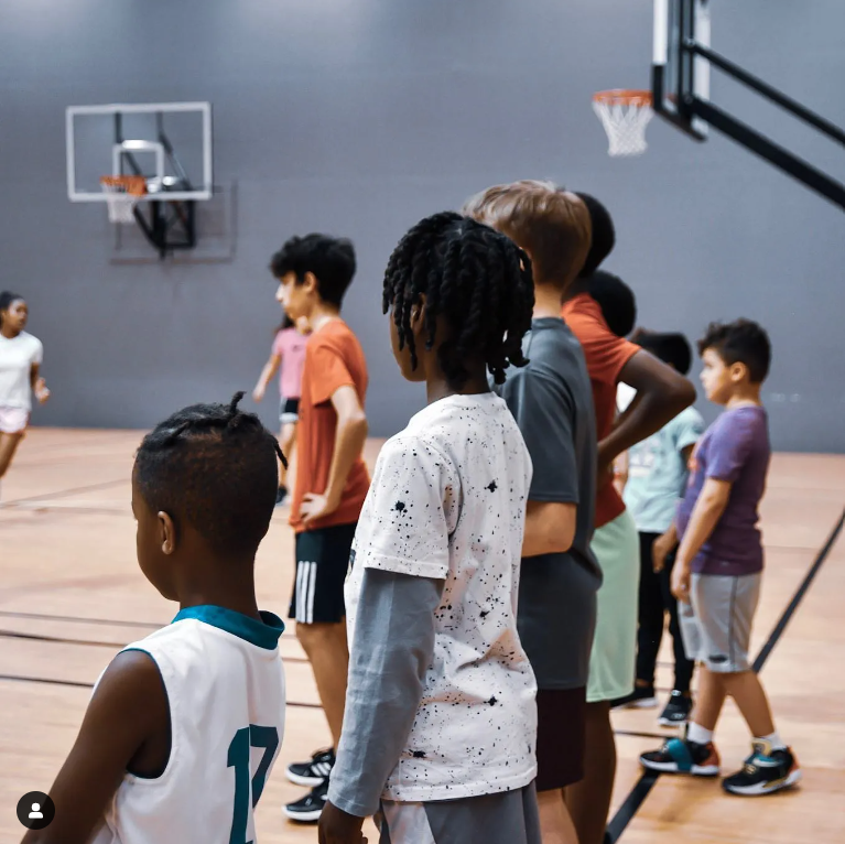Children lined up on a basketball court, watching a basketball game.