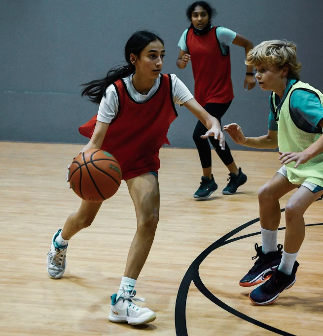 Young woman dribbles a basketball on a court, facing a boy guarding her, two others running behind.