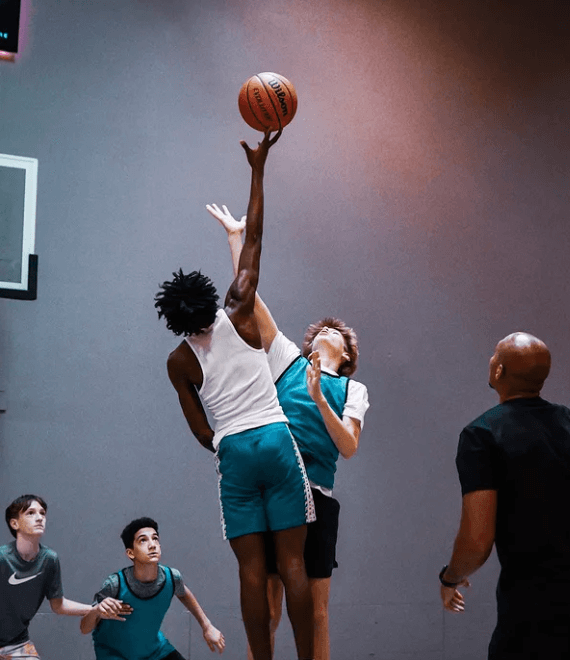 Basketball players jumping for the ball in a gym; one player reaches up, other players look on.