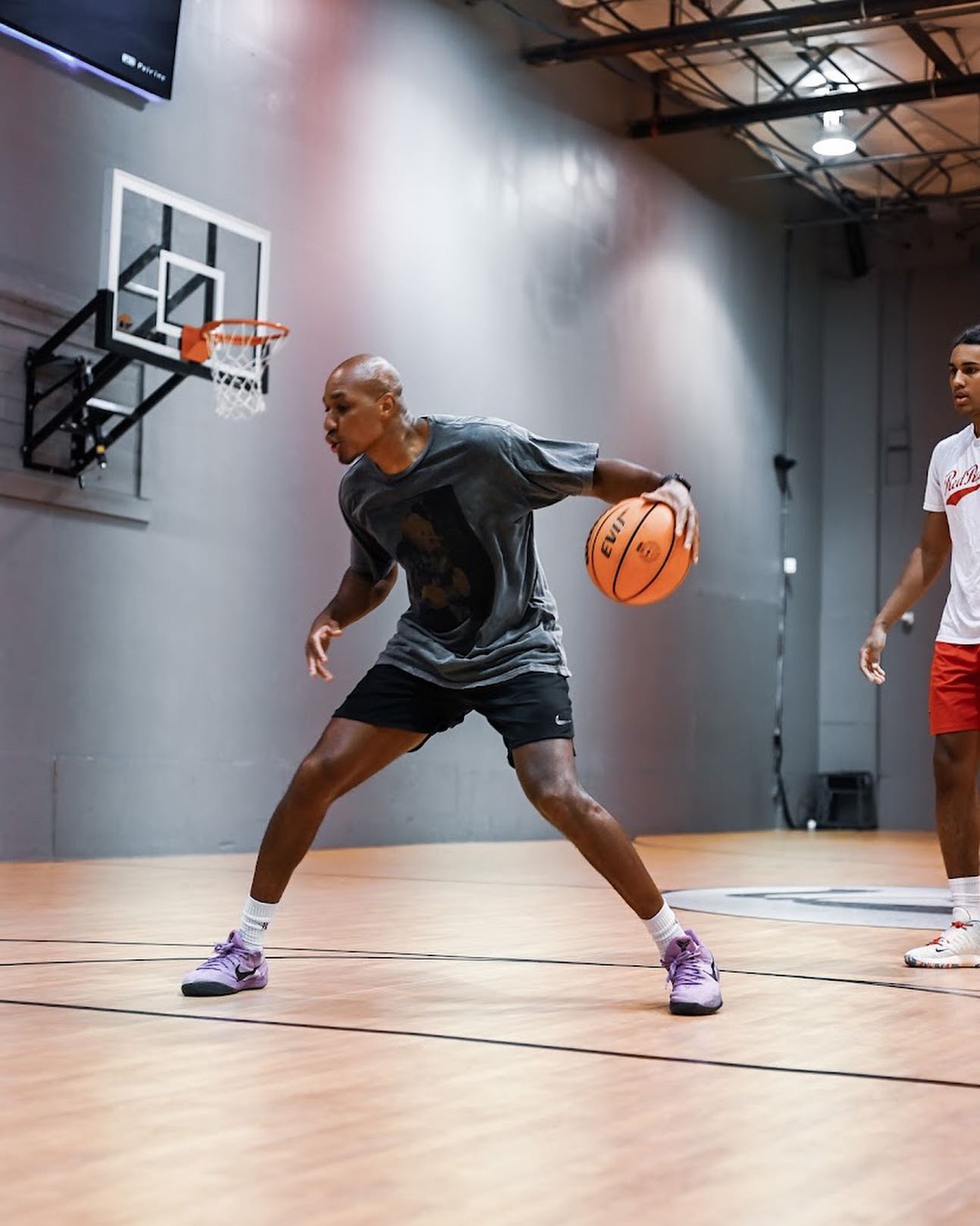 Basketball player dribbles the ball, eyes focused, practicing in an indoor gym. A second player watches.