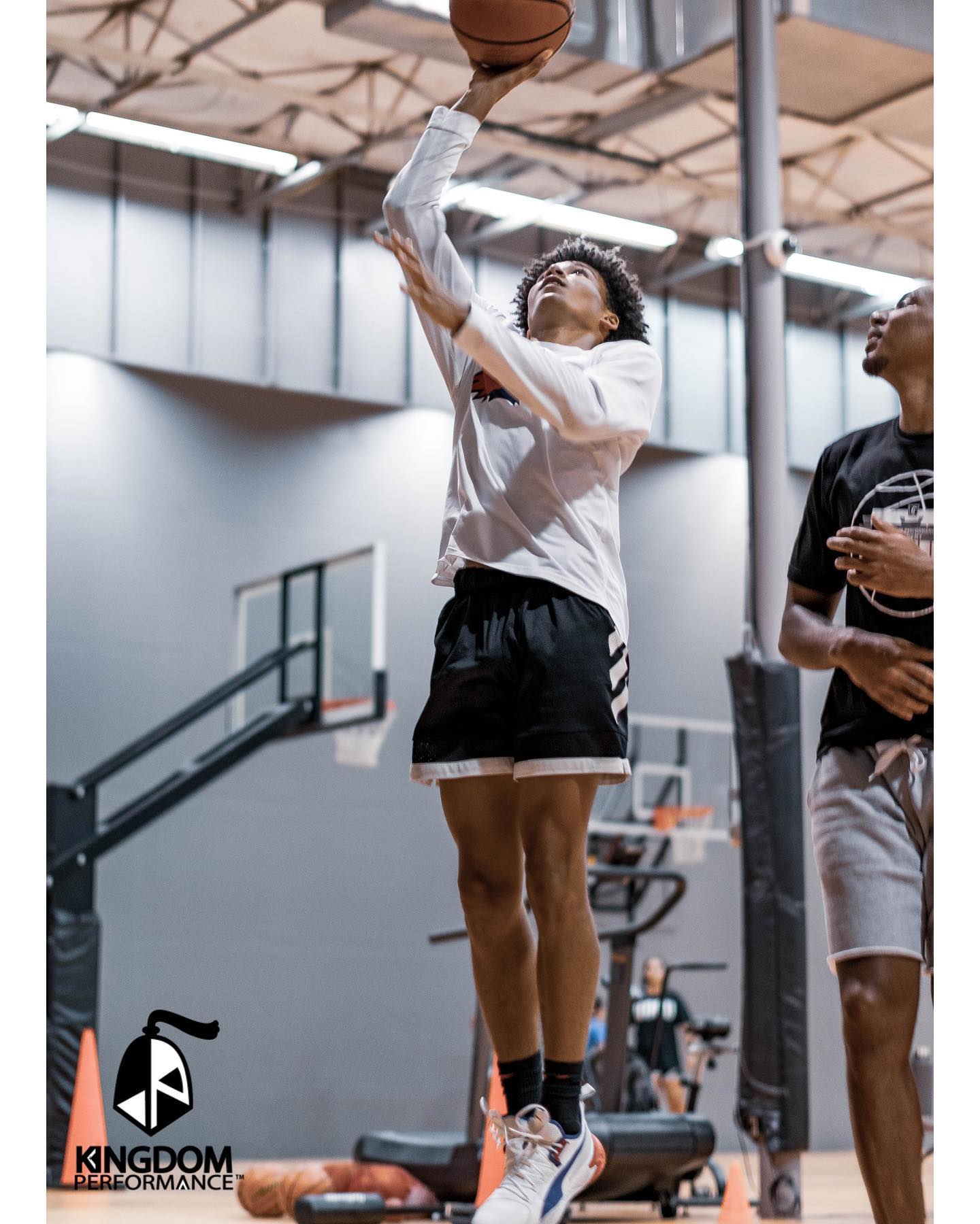Basketball player shooting a ball indoors; white shirt, black shorts, leaping form, another person watching.