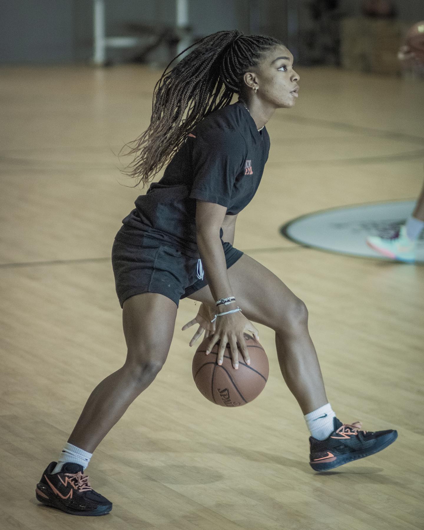 Woman dribbling a basketball on a wooden court, wearing black shorts, shirt, and sneakers.