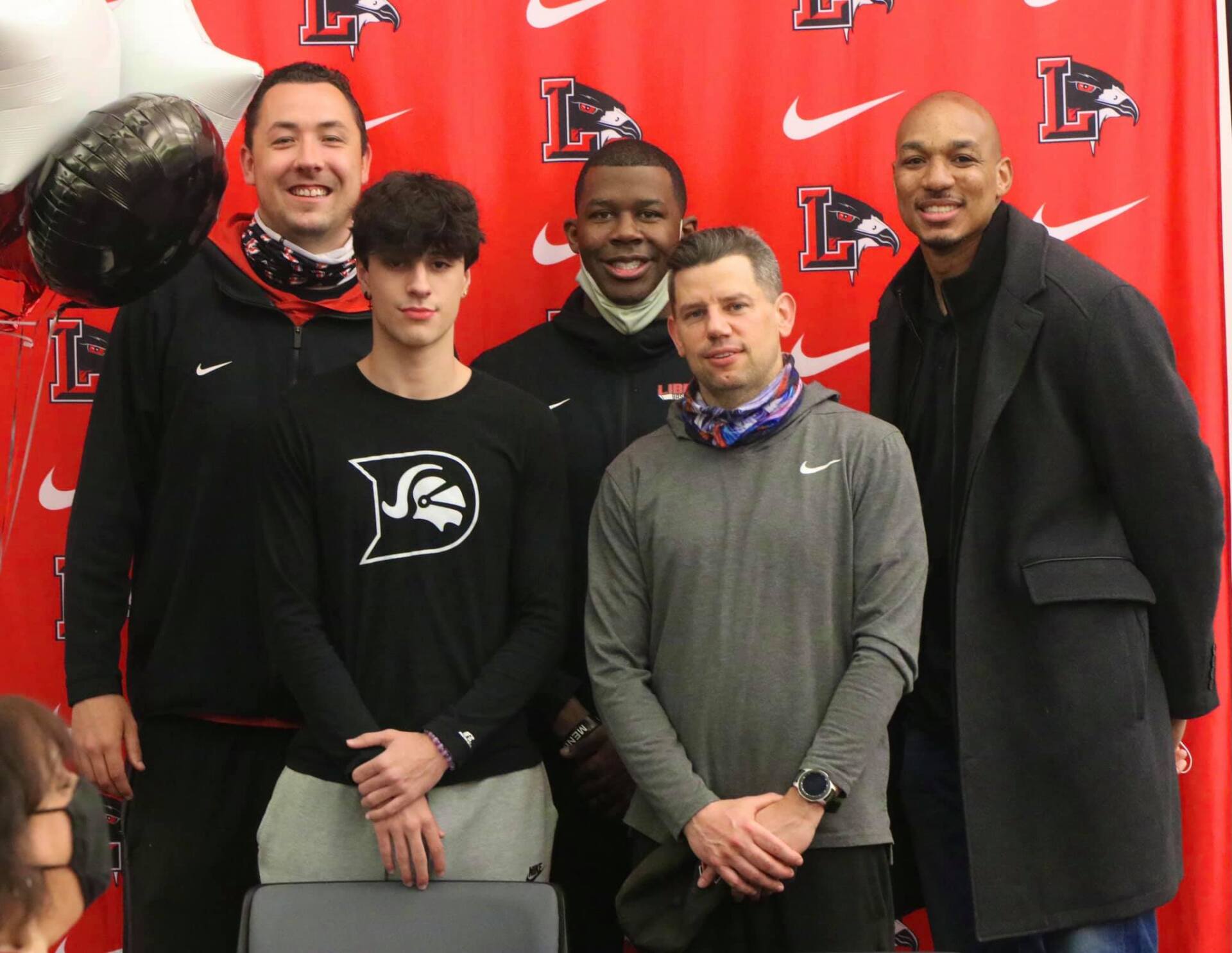 Five people pose in front of a red backdrop. Two young men flank the center, and other three are older. All are smiling.