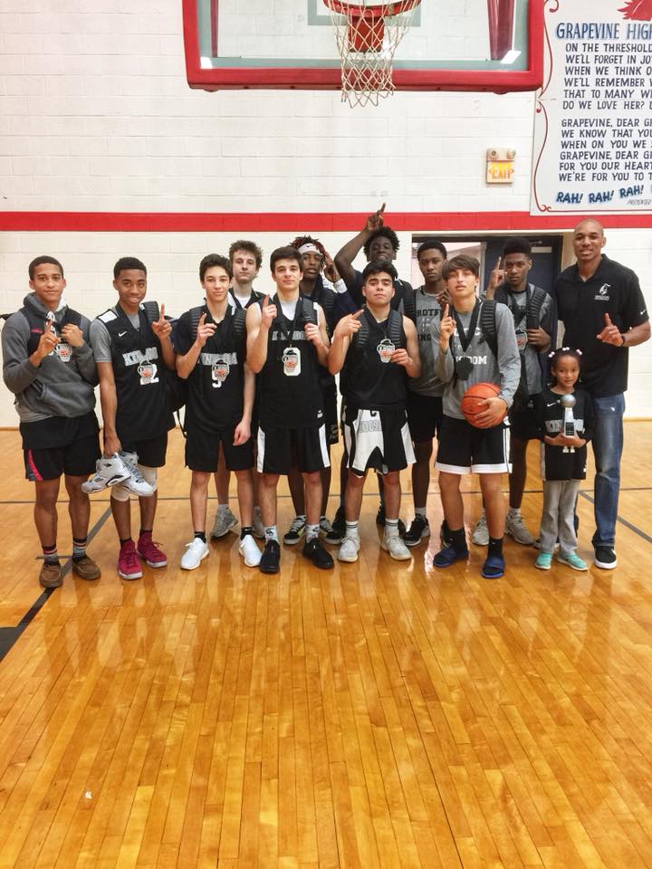 Basketball team poses on court, holding medals. Boys in black jerseys, coach in black polo. Red trim, wooden floor.