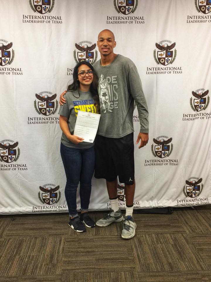 Woman and tall man pose together in front of a banner. The woman holds a paper; both smile.
