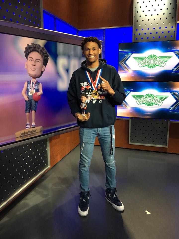 Man in blue hoodie holding trophies, standing next to bobblehead in a studio setting.